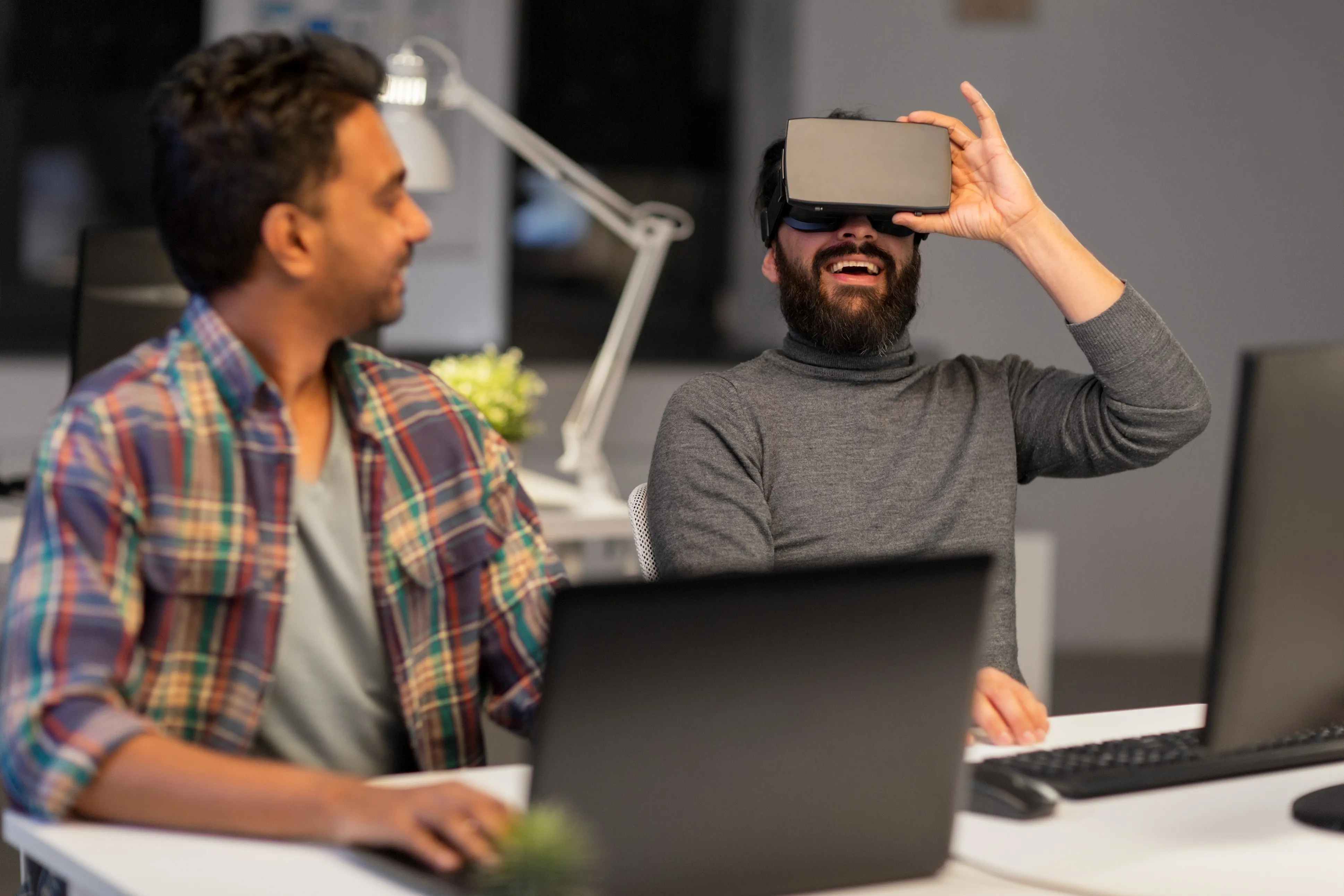 Two engineers at a shared desk, one trying on a VR headset